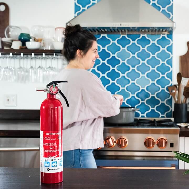 A woman cooks in the kitchen, with a First Alert fire extinguisher on a nearby counter.