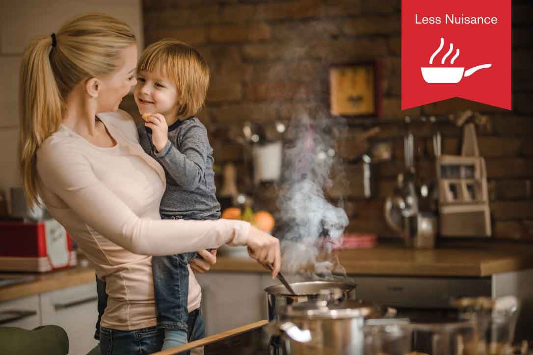 Less Nuisance. A woman holds her young son in her arms while she cooks over the stovetop.